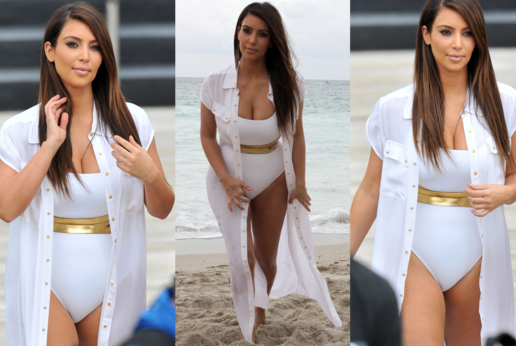 Kim Kardashian i vit baddräkt på stranden. Med guldbälte och lång skjorta. 